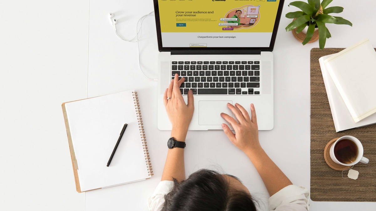 Overhead view of hands typing on a laptop with a webpage open, notebook and coffee on the desk