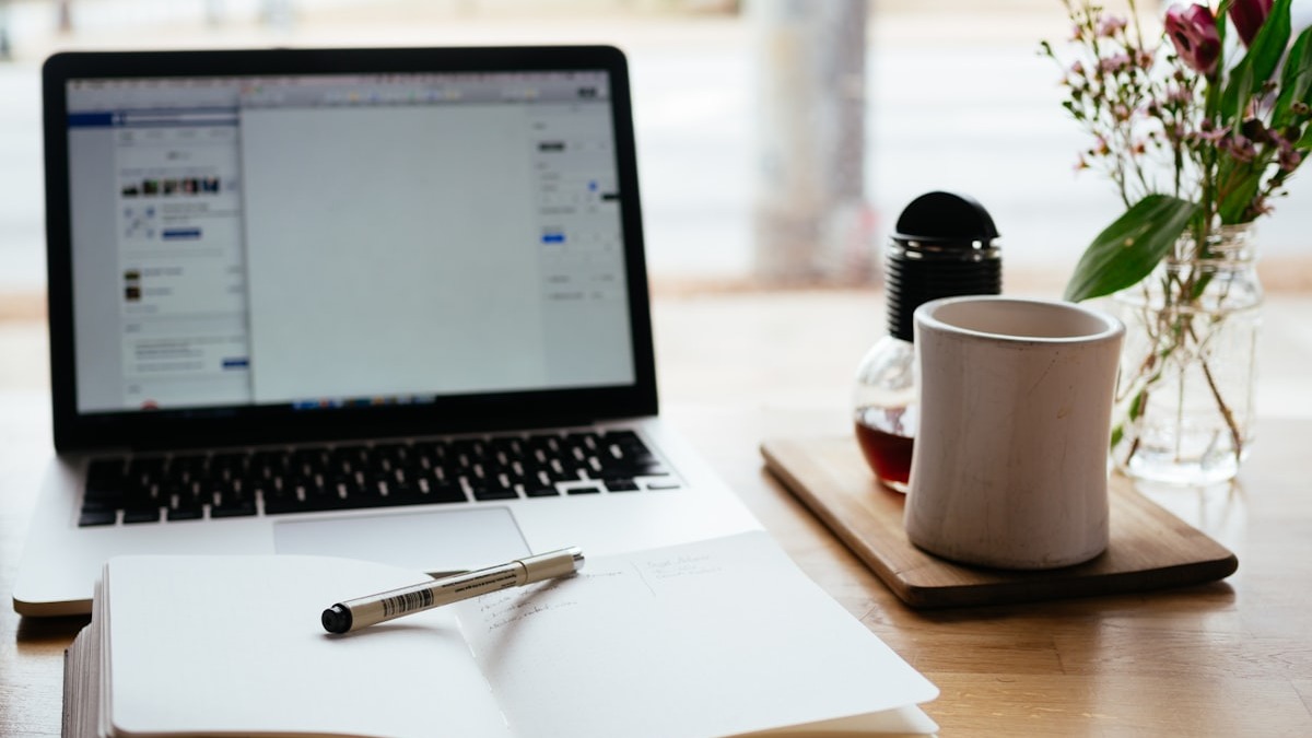 An open notebook with a pen resting on it, beside a laptop, on a wooden desk with coffee