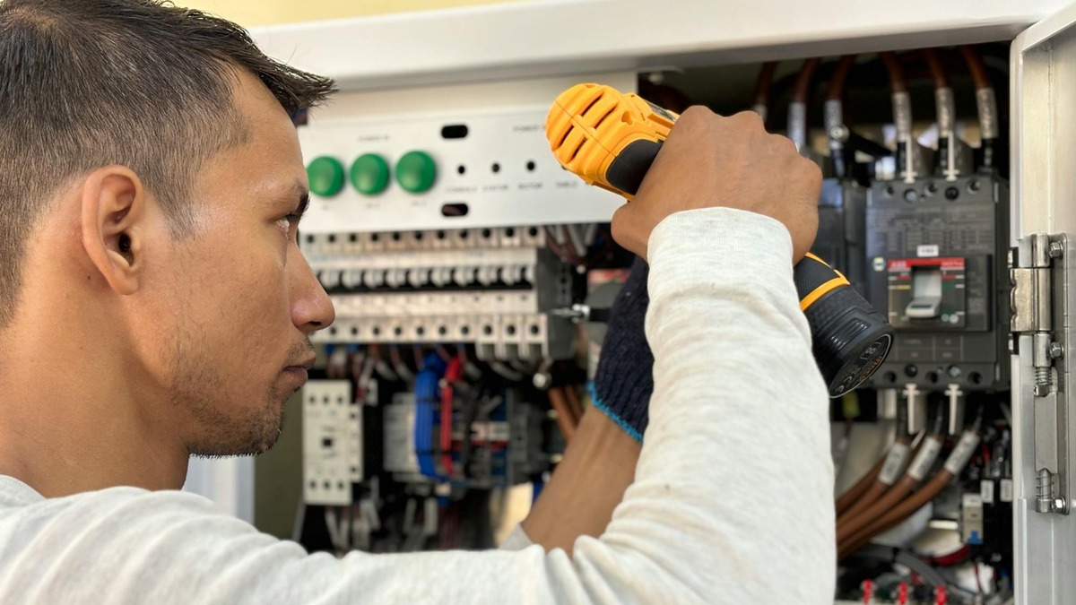 An electrician working on a circuit breaker panel with a power drill