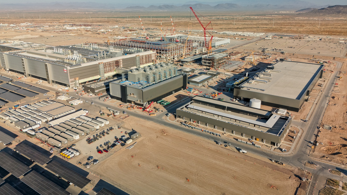Aerial view of a large semiconductor fabrication plant under construction, with cranes and support buildings surrounding the main structure