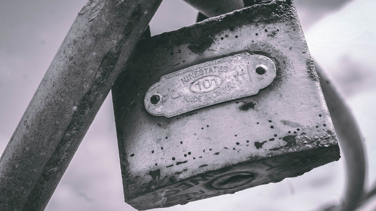 A weathered metal padlock attached to a gray metal bar