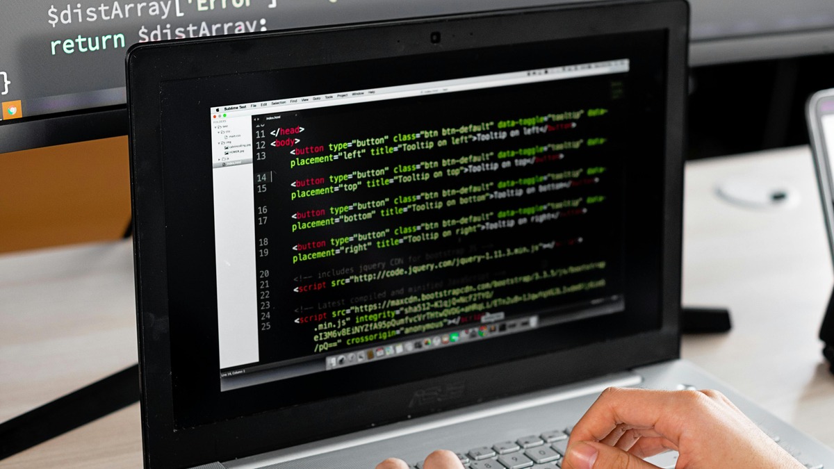 A developer reviewing code on a laptop with an external monitor in a modern office workspace