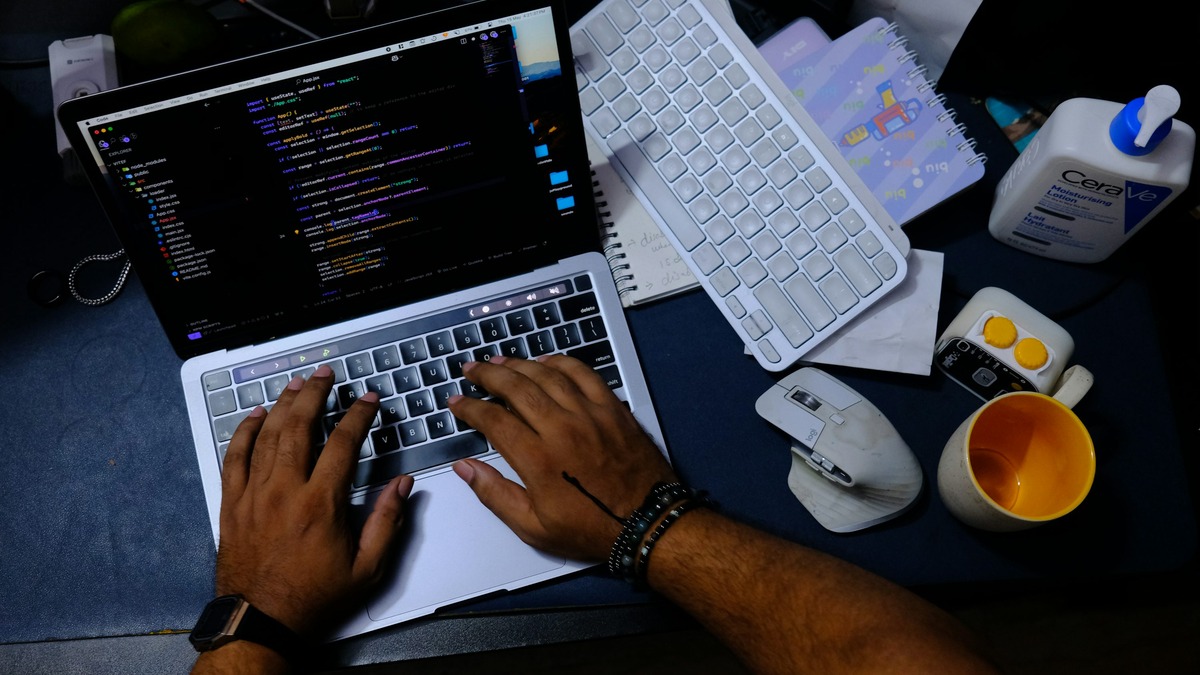 Overhead view of hands typing on a laptop showing code, with a keyboard, mouse, and coffee mug on the desk