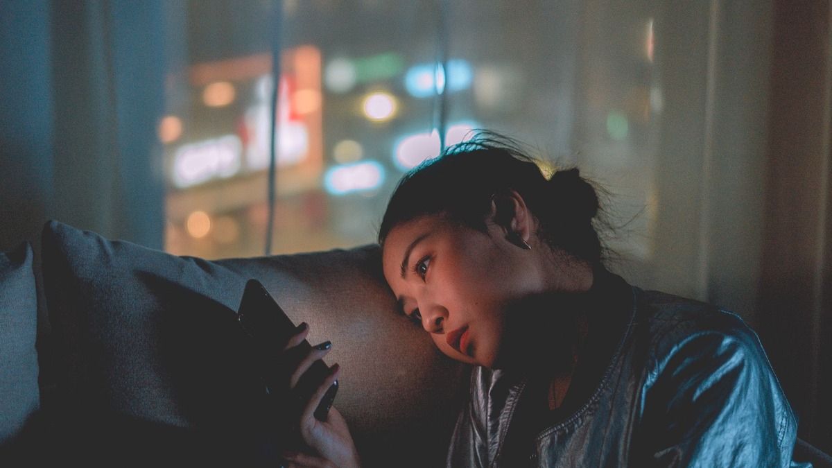 A woman lying on a couch at night, illuminated by her phone screen, looking away toward city lights through the window