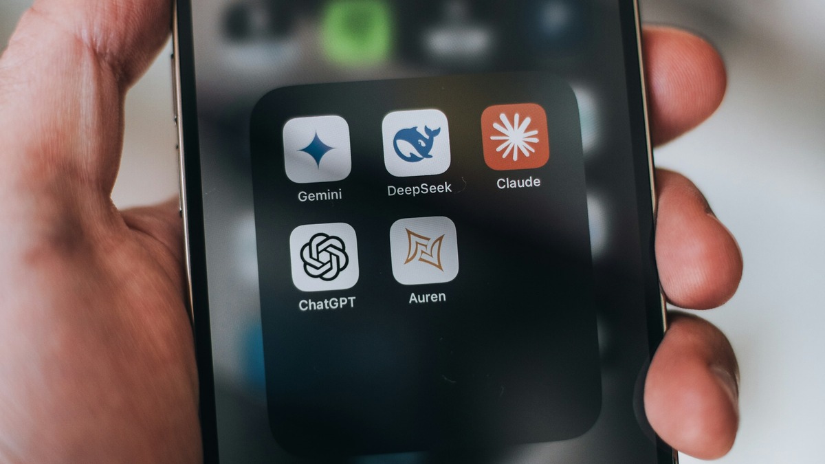 A hand holding a smartphone displaying a folder of AI chatbot apps including Gemini, DeepSeek, Claude, and ChatGPT