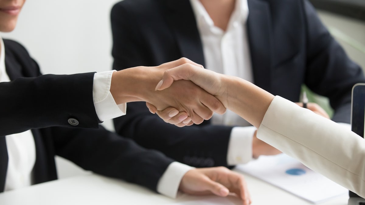 Two people in business attire shake hands across a table with financial documents in view
