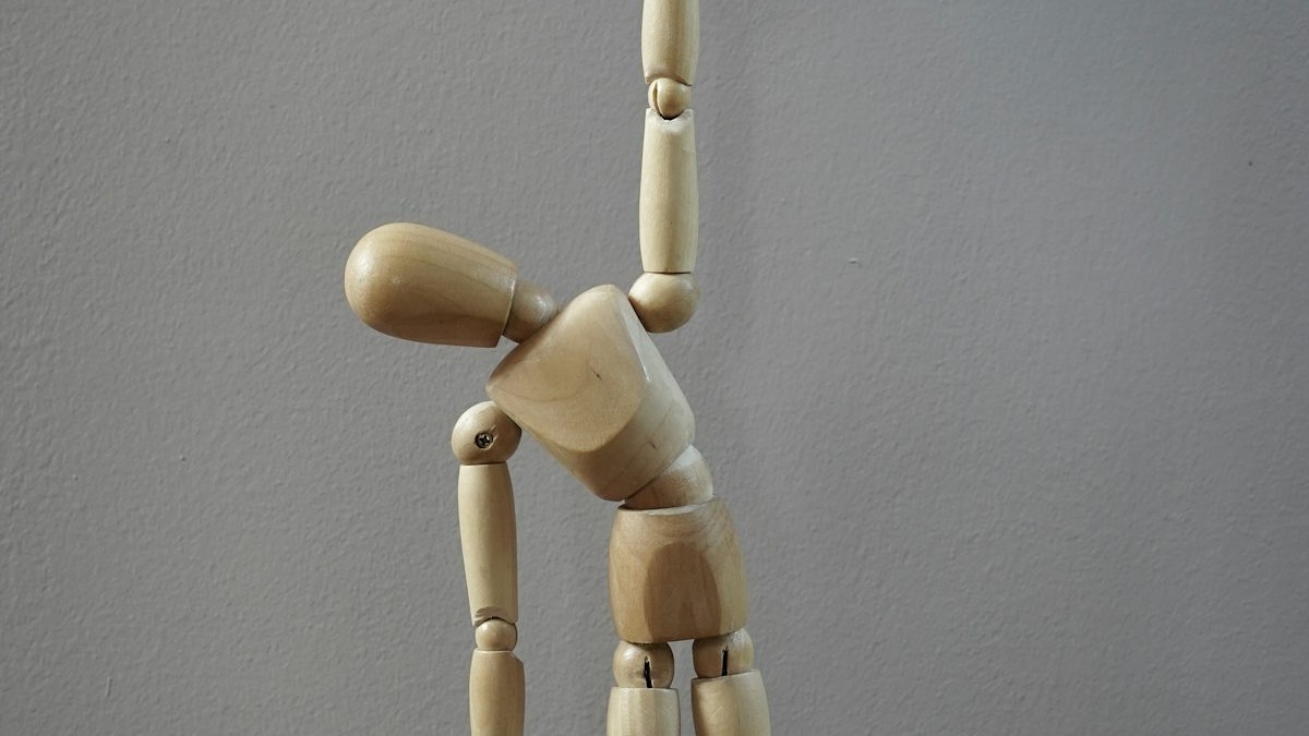 A hand suspends a jointed wooden mannequin figure by a single string against a plain gray background