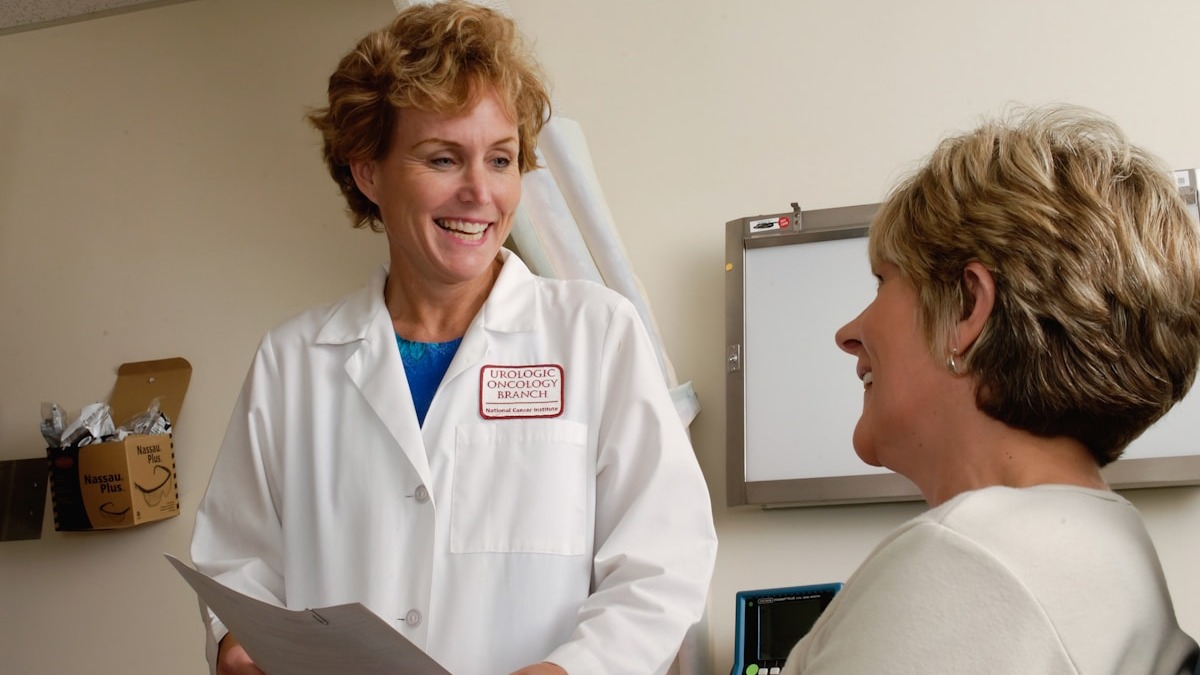A physician consulting with a patient while a laptop displays clinical notes in the background